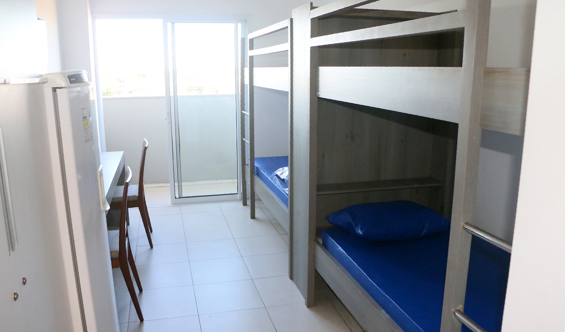 interior de um quarto, mostrando duas camas do tipo beliche, uma geladeira e uma porta que dá para uma varanda